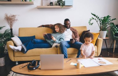 Kafkasyalı eş kitap okurken, bulanık çocuk oynarken sosyal ağlarda sohbet etmek ve mesajlaşmak için cep telefonu cihazını kullanan mutlu koyu tenli adam.