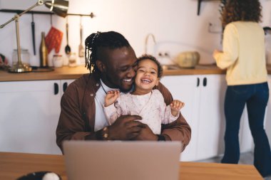 Ev ortamında esnek çalışma ortamı: çocuk bakımı ve uzaktan kumandalı internet cihazını birleştiren ebeveyn, neşeli baba ve komik kızın mutfak çalışma alanında dizüstü bilgisayarla gülmesi