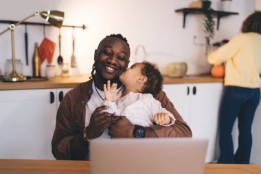 Ev ortamında esnek bir iş yeri: Çocuk bakımı ve uzak işleri modern netbook bilgisayarında birleştiren ebeveyn, neşeli baba gülümserken, çocuk da mutfakta dizüstü bilgisayarla öpüşüyor.