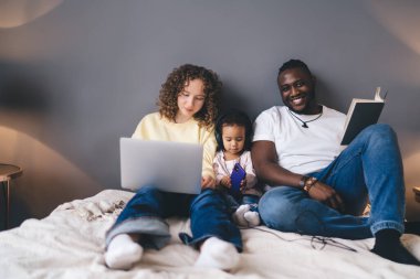 Kıvırcık saçlı anne dizüstü bilgisayar kullanırken baba kameraya gülümserken kulaklıklı küçük çocukları ile rahat evlerinin iç kısımlarına teknolojiyi sorunsuz bir şekilde entegre ediyor.