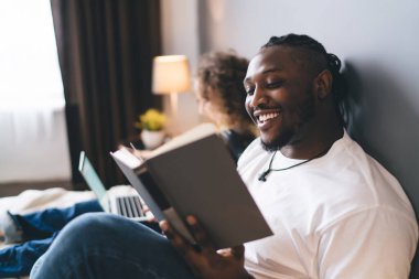 Neşeli Afro-Amerikalı adam, boş zamanlarında eğleniyor ve evde ilginç bir kitap okuyor, neşeli esmer tenli hippi elinde edebiyat şiiri tutuyor ve hobi sırasında gülümsüyor.