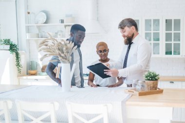 Profesyonel sakallı, resmi kıyafetli şişman adam modern güneşli dairede mutfak masasının yanında duran ilgili etnik çifte evrak gösteriyor.