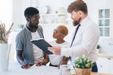 Genç Afrikalı evli çift birbirlerine bakarken emlakçı beyaz mutfağın arka planında daire almayı öneriyor.