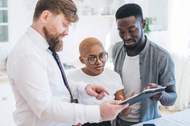 Konsantre olmuş Afro-Amerikan evli çift emlakçıyı dinliyor. Apartman kontratı öneriyor. Beyaz mutfakta duran belgelerle panoyu işaret ediyor.