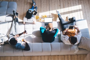 Aydınlık bir dairede kanepeye oturmuş çeşitli cihazlar kullanarak ve sosyal medyayı karıştırarak dinlenen çok ırklı arkadaş grubundan.