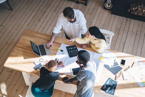 From above diverse business team sitting and standing around table and analyzing data on laptop while working in light office together