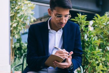 Yakışıklı, şık giyimli ve gözlüklü erkek girişimci. Kafe terasında uzaktan çalışırken notlar alıyor. Kendine güveni tam, zengin bir blog yazarı.