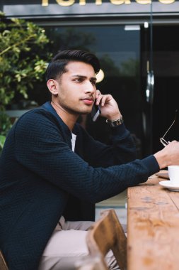 Ciddi müreffeh Hindu milenyum erkek serbest çalışan cep telefonuyla iş konuşan yetenekli bir adam bilgisayarla kafeterya terasında çalışıyor ekran araması yapıyor.