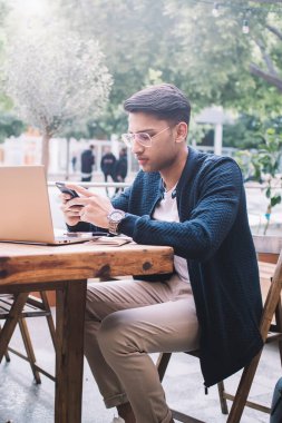 Kendine güvenen erkek CEO laptopta uzaktan çalışıyor. Sosyal ağlarda haberleri akıllı telefondan kontrol ediyor. Yakışıklı Hindu iş adamı dışarıda dizüstü bilgisayarla serbest çalışıyor.