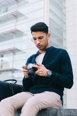 Ciddi Hintli erkek fotoğrafçı modaya uygun giyiniyor ve gözlük takıyor. Profesyonel ekipmanları kontrol ediyor. Düşünceli gezgin şehir merkezinin fotoğraflarını çekmek için kamera kullanıyor.
