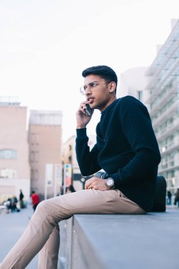 Ciddi Hindu erkek modaya uygun giyinmiş ve gözlüklü. Şehir merkezinde meydanda oturmuş cep telefonuyla konuşuyor. Gezgin, dalgın hipster adam.