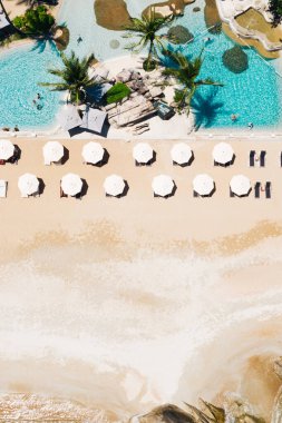 Açık havuzu ve güneş pansiyonları olan ilk hat plaj otelinin havadan görünüşü. Yaz tatillerinin kuş bakışı manzarası, turkuaz deniz suyu ve temiz kum kıyısındaki şemsiyeler.