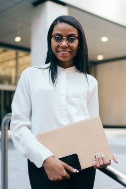 Formal giyim klasör ve akıllı telefon tutarak kameraya gülümseyen çalışan yarım uzunluk portre müreffeh siyah tenli. Ofis Binası yakınında duran başarılı Afro-Amerikan işkadını