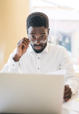 Optik gözlüklü afro-amerikan erkek girişimci muhasebe belgeleri ve finansal web sitesinden gelen bilgileri kontrol etmek için göz korumasını sağladığı için şoke oldu.