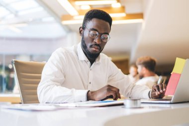 Görme düzeltmesi için göz giysisi giyen konsantre Afro-Amerikan erkek çalışanı. Finans raporlarını okuyor ve analiz ediyor. Ciddi, koyu tenli bir erkek yönetici, dizüstü bilgisayarla çalışma masasında oturuyor.