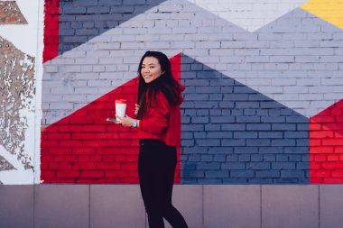 Renkli şehirlerde poz vermek için kahveyle gezen neşeli Asyalı genç kadın portresi, şık giyinmiş komik Japon milenyumu sokak sanatıyla turistik bölgeyi ziyaret ediyor.