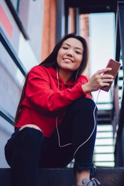 Kulaklıklı neşeli hipster kız portresi radyo yayını dinliyor ve şehir merdivenlerinde dinlenen kameraya gülümsüyor, kulaklıklı mutlu Japon kullanıcı boş zamanlarında ses şarkısının keyfini çıkarıyor.