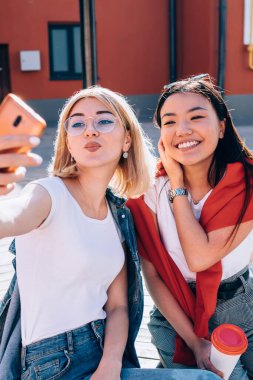 Şehir ortamında arkadaşça toplanan neşeli, samimi kadınlar, eğlence ortamında komik içerik fotoğrafları çekmek için poz veren neşeli binyıllar boş zamanlarında ön telefon kamerası kullanan kadınlar.