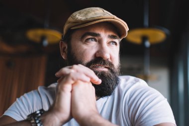 Ciddi sakallı, modaya uygun bir şapkası ve günlük kıyafetleri olan bir adam kafeteryada oturuyor. Elleri yüzüne yakın ve düşünceli bir şekilde başka tarafa bakıyor.