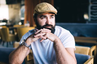 Düşünceli, siyah sakallı bir adam boş bir kafede oturuyor. Masasında defter ve kahve var ve dalgın dalgın bakıyor.