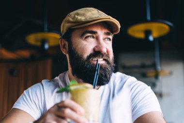 Sakallı, gülümseyen ve soğuk bir içecek içen erkek. Tatil sırasında defter ile çalışırken şık, modern, karanlık bir kafeye bakıyor.