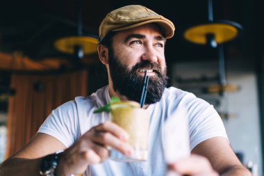 Sakallı, gülümseyen ve soğuk bir içecek içen erkek. Tatil sırasında defter ile çalışırken şık, modern, karanlık bir kafeye bakıyor.