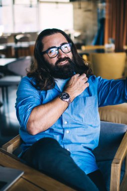 Sıradan giyinmiş, gözlüklü, uzun saçlı zeki genç bir adam koltukta oturuyor ve kafede akıllı telefondan selfie çekiyor.