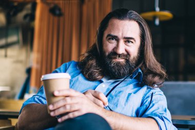 Pozitif sakallı genç adam, uzun saçlı, gülümseyen ve kameraya bakan iş sırasında masanın yanındaki kafede dizüstü bilgisayarla oturuyor.