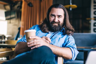 Pozitif sakallı genç adam, uzun saçlı, gülümseyen ve kameraya bakan iş sırasında masanın yanındaki kafede dizüstü bilgisayarla oturuyor.