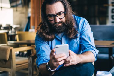Ciddi sakallı, olgun, serbest çalışan uzaktan kumandalı telefondan mesajları ve sosyal ağları kontrol eden 40 yaşında erkek, cep telefonu uygulaması üzerinden işlem yapıyor.