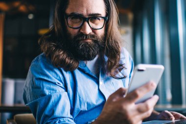 Akıllı telefon tutan yakışıklı erkek girişimcinin portresi ve uzaktan kumanda sırasında kameraya bakmak, olgun 40 'lı yaşlarda serbest çalışan bir adam bilgisayarla kafenin içini tarıyor ve paylaşıyor.