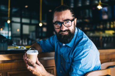 Günlük giysiler içinde sakallı bir erkek, bir bardak sıcak içecekle birlikte, moda barın ahşap tezgahında oturuyor ve başka tarafa bakıyor.