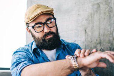 Gözlüklü, gözlüklü, sıradan kıyafetleri olan ve kafede otururken kol saatine bakan modaya uygun yetişkin bir hippi.