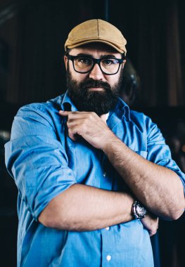 Kot gömlekli ve kepli hippi bir adam karanlık iç mekanda dikilirken kameraya ciddi ciddi bakıyor.