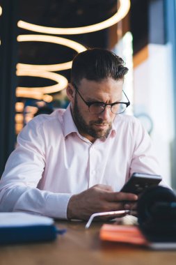 Optik göz giyimli genç serbest çalışan masa başına oturmuş uzaktan kumandalı iş sırasında akıllı telefon tebligatı kuruyor, genç bir erkek cep telefonuyla ilgili bilgi arıyor