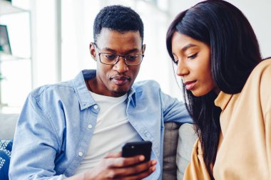 Afro-Amerikalı bir adam evde rahat bir oturma odasında kanepede otururken etnik kız arkadaşıyla akıllı telefondan bilgi paylaşıyor.