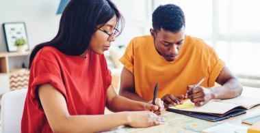 Konsantre olmuş çok ırklı üniversite öğrencileri, aydınlık odada kağıt ve dizüstü bilgisayarlarla masaya otururken proje üzerinde birlikte çalışıp notlar alıyorlar.