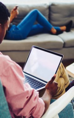 Yukardan bakıldığında sıradan giyinen siyahi erkek girişimci, şık oturma odasındaki koltukta otururken boş ekran dizüstü bilgisayar kullanıyor.