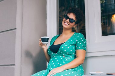 Heyecanlı gülümseyen, sıradan giyinmiş, güneş gözlüklü bir kadın bir fincan sıcak içecek içiyor ve sokak kafesinde eğleniyor.