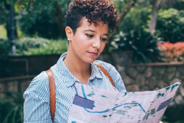 Sakin, kıvırcık saçlı, sırt çantalı, kağıt haritaya bakan ve tropikal parkta gezinen genç bir kadın.