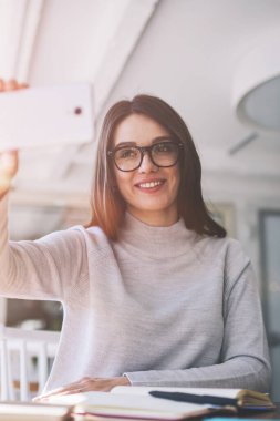 Cep telefonu kamerasına bakıp kendini gösteren mutlu kadın blogcu, kafeteryada oturan başarılı hippi kız ve selfie çekmek için akıllı telefonu kullanıyor.