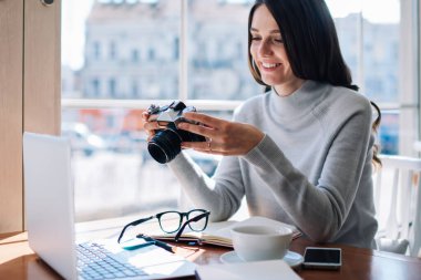 Pozitif kadın fotoğrafçı elinde retro kamerayla kahve dükkanında oturup görüntüleri kontrol ediyor. En sevdiği hobi sırasında eğlenen başarılı amatör hippi kız.