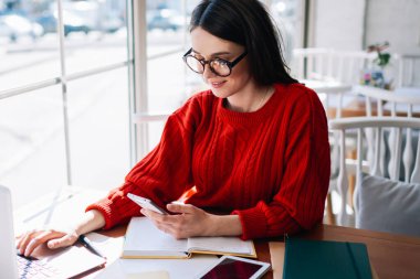 Pozitif kadın yazılım geliştirici kafeteryadaki dizüstü bilgisayarda çalışırken cep telefonundan mesajları kontrol ediyor, kapalı mekanlarda kablosuz internete bağlı sosyal ağlarda son derece mutlu hipster kız sohbeti yapıyor.