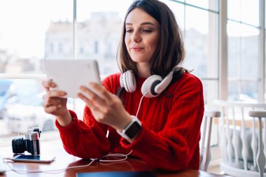 Dijital tablette oyun oynayan konsantre hipster kafeteryada otururken ve kablosuz internet alanına bağlanırken, mutlu genç kız arkadaşıyla taşınabilir bilgisayar kullanarak video sohbeti yapıyor.