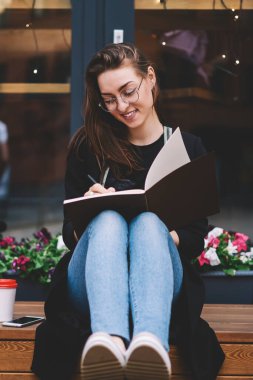 Reklam alanında oturup gülümseyen, neşeli hippi kız ders çalışmaktan mutlu olurken heyecanlı bir fikirle kompozisyon yazan pozitif kadın yazar ders kitabında
