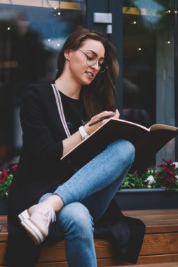 Üniversite kampüsünde eğitim için ders kitabı tutan ve ödev yapmaya hazırlanan çekici, zeki hippi kız, gözlüklü çekici kadın üniversitede yedek kulübesinde proje planlıyor.