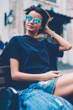 Modaya uygun hippi kız şık gözlük takar ve şık kot pantolon giyer. Bankta dinlenirken ve beklerken yüzünü çevirir. Pozitif bayan gezgin şehir sokaklarında tatilin tadını çıkarır.