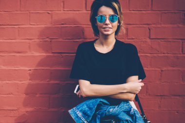 Kot pantolon ve şık güneş gözlüğü takan 20 'li yaşlardaki neşeli beyaz modellerin yarım boy portresi akıllı telefon tutuyor ve reklam mesajları ve logolar için fotokopi alanının yanında duruyor.