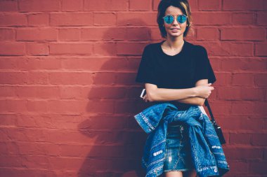 Kot pantolon ve şık güneş gözlüğü takan 20 'li yaşlardaki neşeli beyaz modellerin yarım boy portresi akıllı telefon tutuyor ve reklam mesajları ve logolar için fotokopi alanının yanında duruyor.