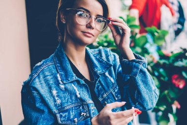 Düşünen bir kadın şık kot ceket giyer ve şık gözlükler takar. Düşünceli bir şekilde başka yere bakar, rahat bir sokak kafesinde eğlenir ve bekler. Hipster yaşam tarzı ve dinlenme konsepti.